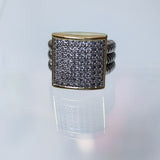 A square face ring featuring a gold-toned bezel-set pavé band with clear stones, flanked by twisted silver-toned side bands, displayed on a reflective white surface.