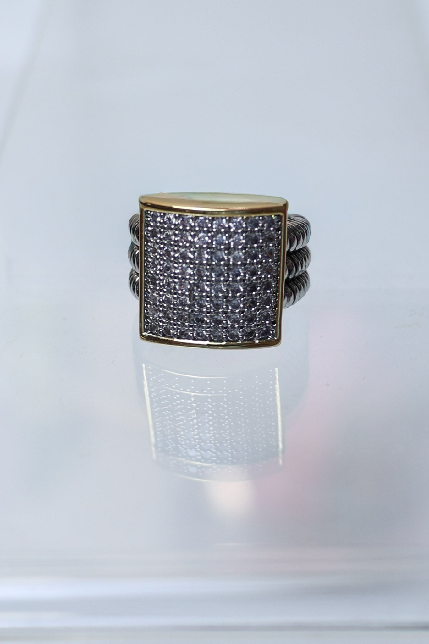 A square face ring featuring a gold-toned bezel-set pavé band with clear stones, flanked by twisted silver-toned side bands, displayed on a reflective white surface.