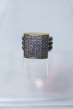 A square face ring featuring a gold-toned bezel-set pavé band with clear stones, flanked by twisted silver-toned side bands, displayed on a reflective white surface.