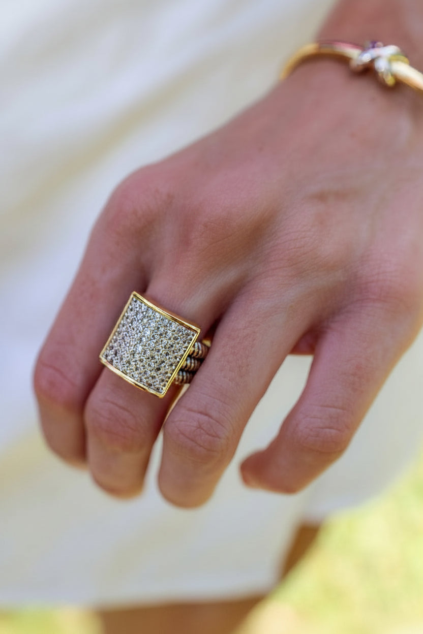 A close-up of a hand wearing a large gold signet ring with a square face paved with small clear stones, alongside a delicate gold bracelet on the wrist.