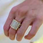 A close-up of a hand wearing a large gold signet ring with a square face paved with small clear stones, alongside a delicate gold bracelet on the wrist.