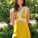 A woman wearing a sleeveless mustard yellow dress with cream lace trim, standing outdoors in front of green foliage.