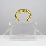 A clear transparent handbag with a glossy gold top handle, gold hardware, and a matching clear detachable shoulder strap, photographed against a black and white studio background.