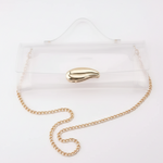 Clear acrylic clutch bag with a gold leaf-shaped clasp and a gold chain strap, shown on a white surface with a black background.