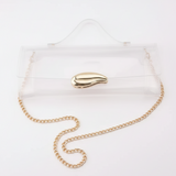 Clear acrylic clutch bag with a gold leaf-shaped clasp and a gold chain strap, shown on a white surface with a black background.