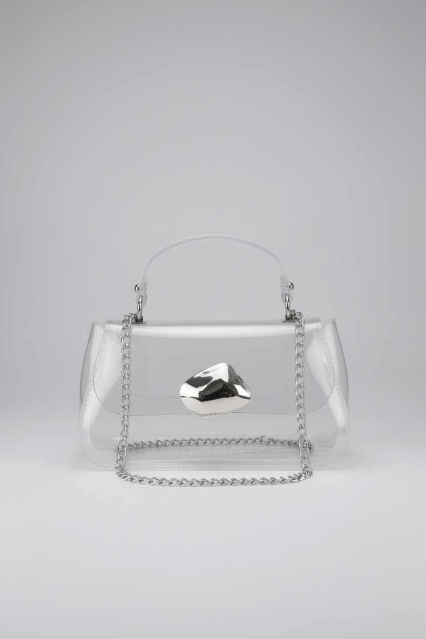A clear acrylic handbag with a silver chain handle and a faceted silver metal accent on the front, photographed against a neutral light gray background.