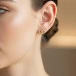 A close-up of a woman's ear wearing a tiny gold hoop earring with a central round cluster of clear stones, set against a softly blurred warm beige background.