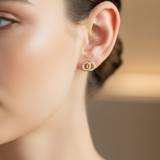 A close-up of a woman's ear wearing a tiny gold hoop earring with a central round cluster of clear stones, set against a softly blurred warm beige background.