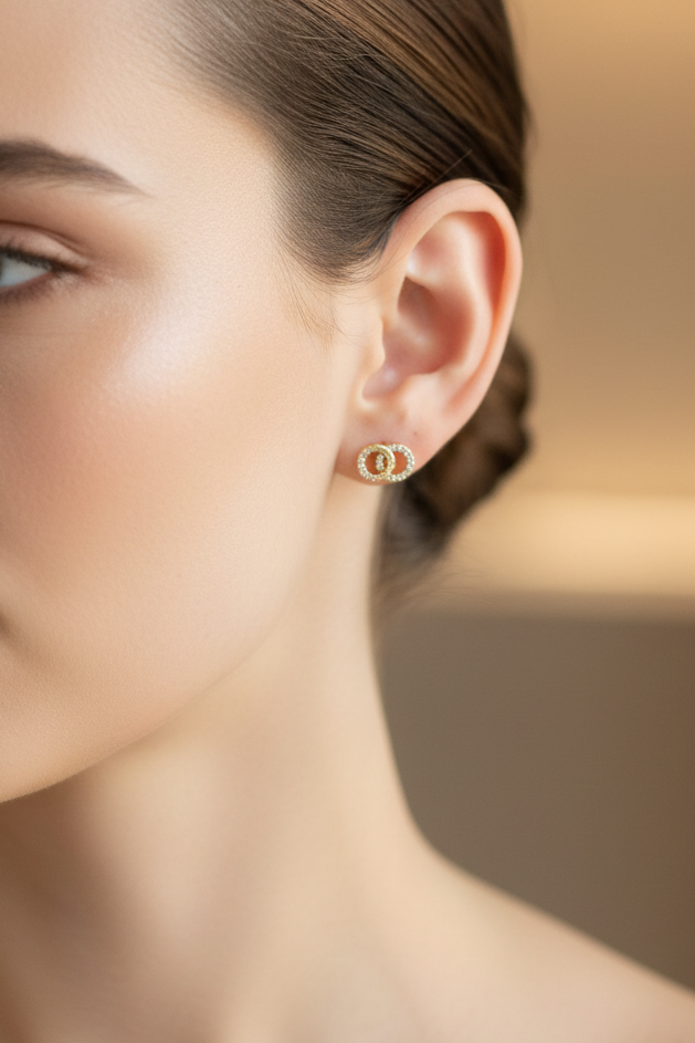 A close-up of a woman's ear wearing a tiny gold hoop earring with a central round cluster of clear stones, set against a softly blurred warm beige background.