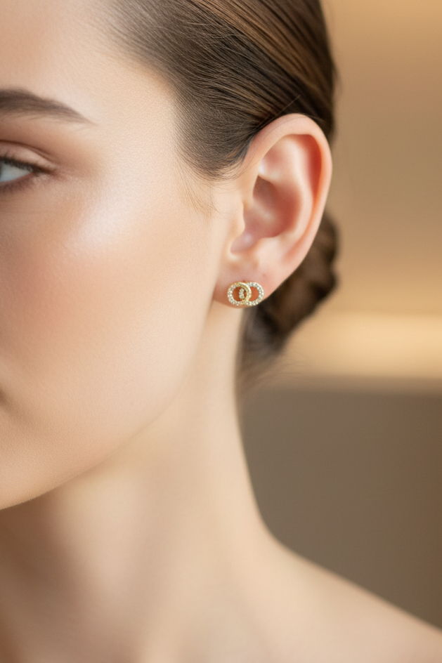A close-up of a woman's ear wearing a tiny gold hoop earring with a central round cluster of clear stones, set against a softly blurred warm beige background.