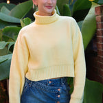 Person wearing a yellow sweater and blue denim skirt standing in front of green plants.