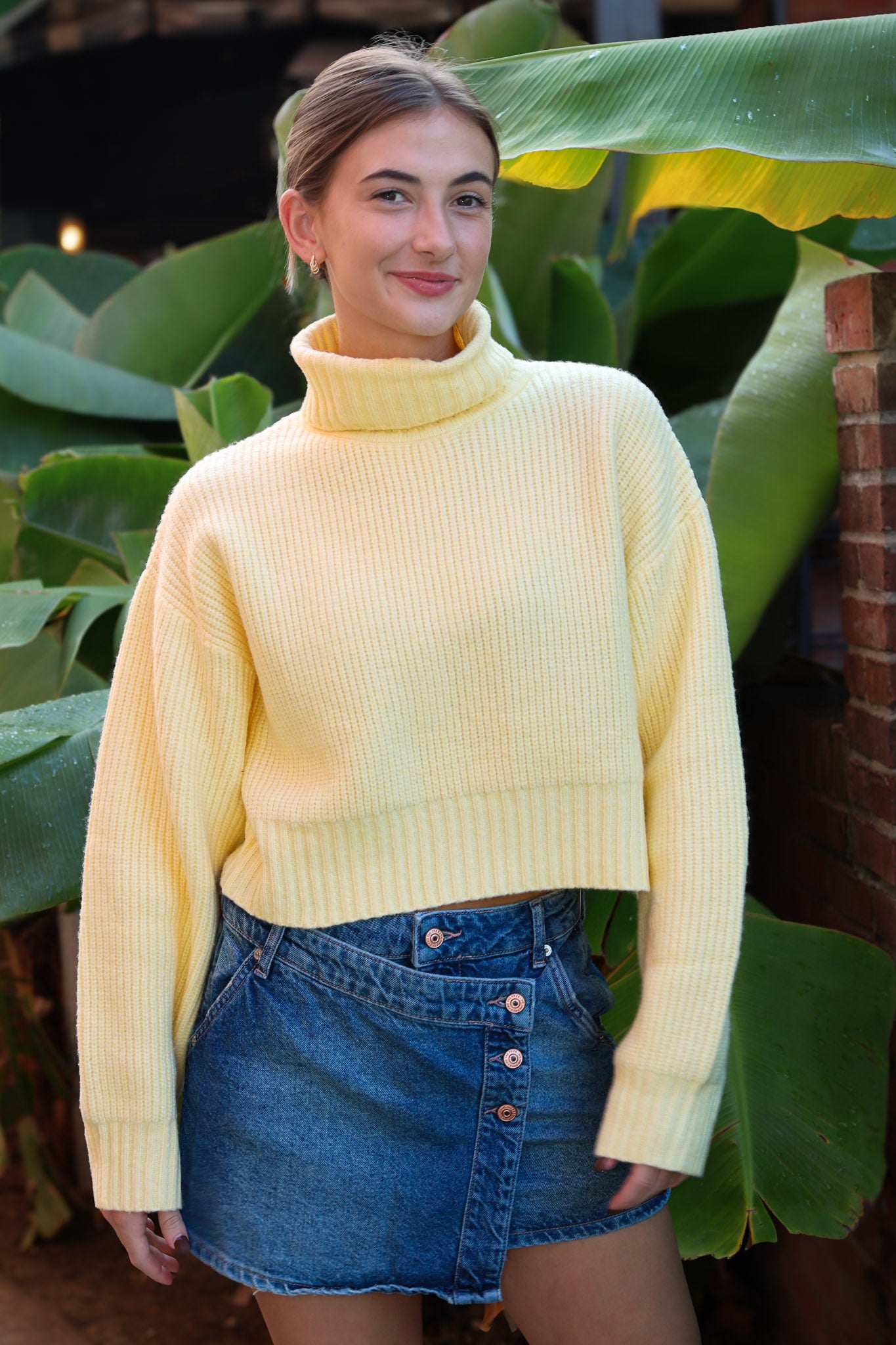 Person wearing a yellow sweater and blue denim skirt standing in front of green plants.