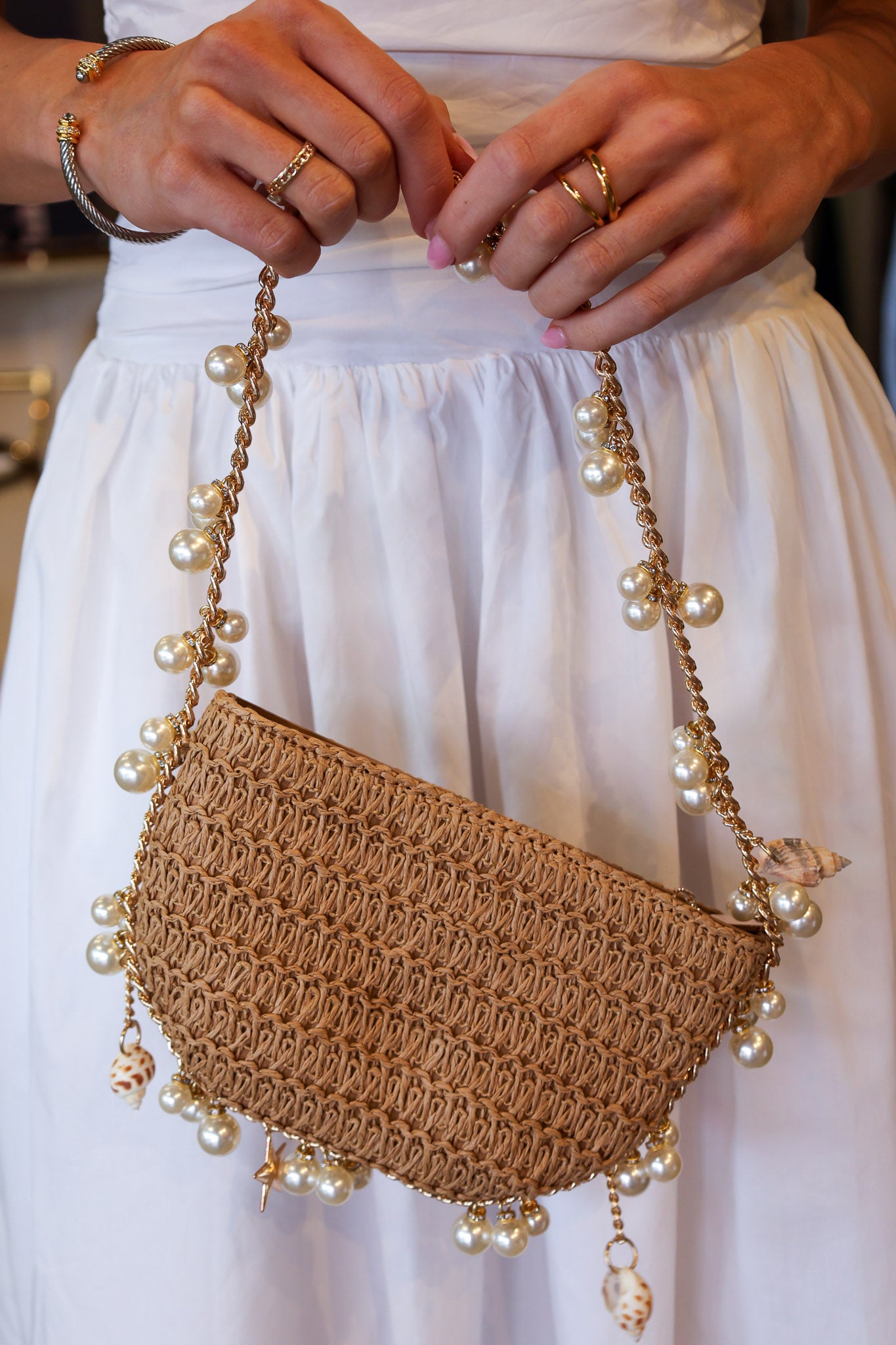 Brown woven handbag with pearl embellishments held by a person wearing a white dress.