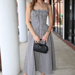 Woman in a checkered dress holding a black handbag on a sidewalk.