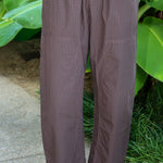 A person wearing loose brown gingham drawstring pants by PISTOLA with a small-scale check pattern, standing outdoors on a concrete surface with green plants in the background.