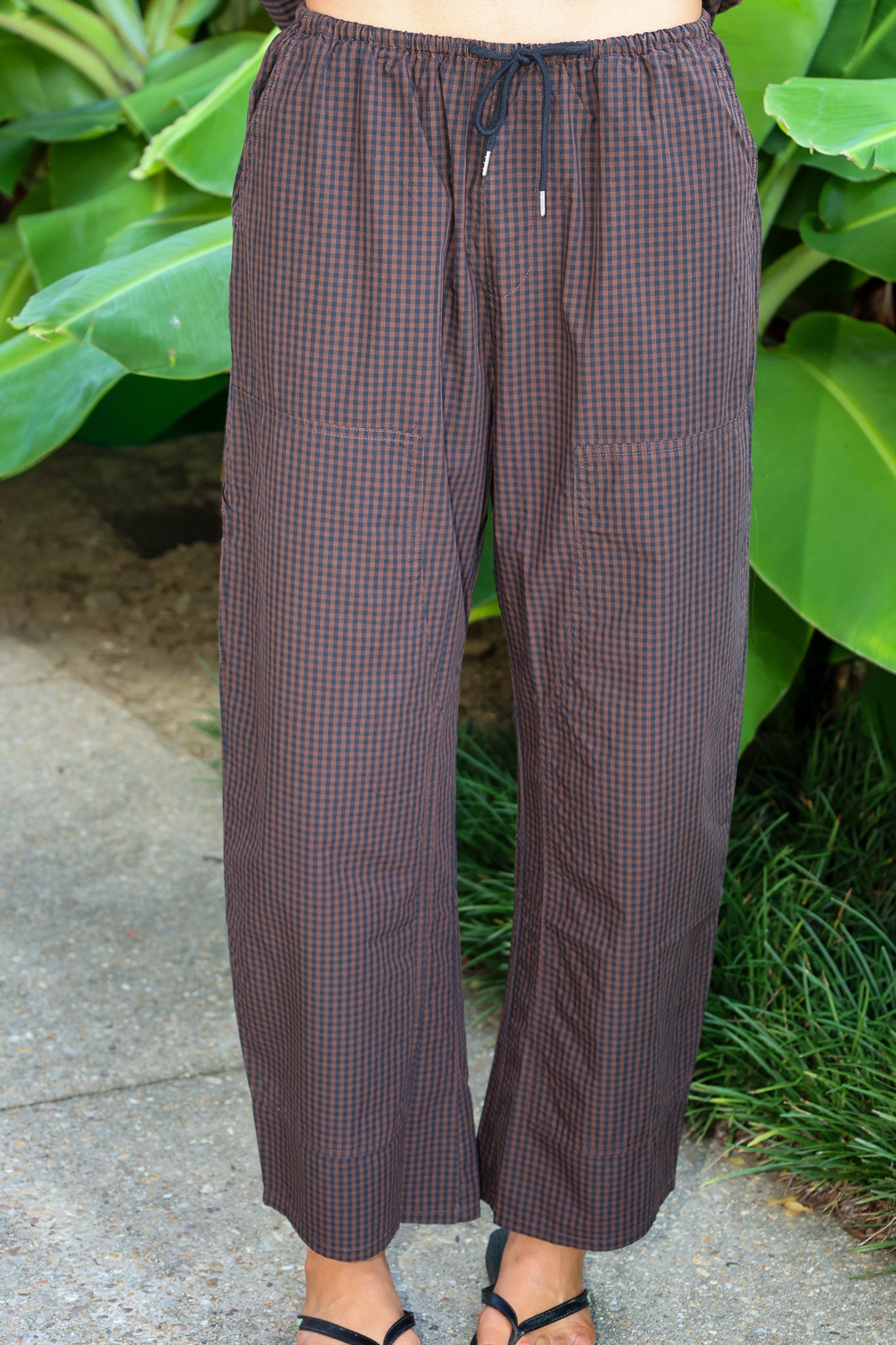 A person wearing loose brown gingham drawstring pants by PISTOLA with a small-scale check pattern, standing outdoors on a concrete surface with green plants in the background.