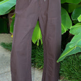 A person wearing loose, wide-leg cropped barrel pants by PISTOLA in a dark brown and black micro-check pattern, standing outdoors on a stone surface with green tropical leaves in the background.