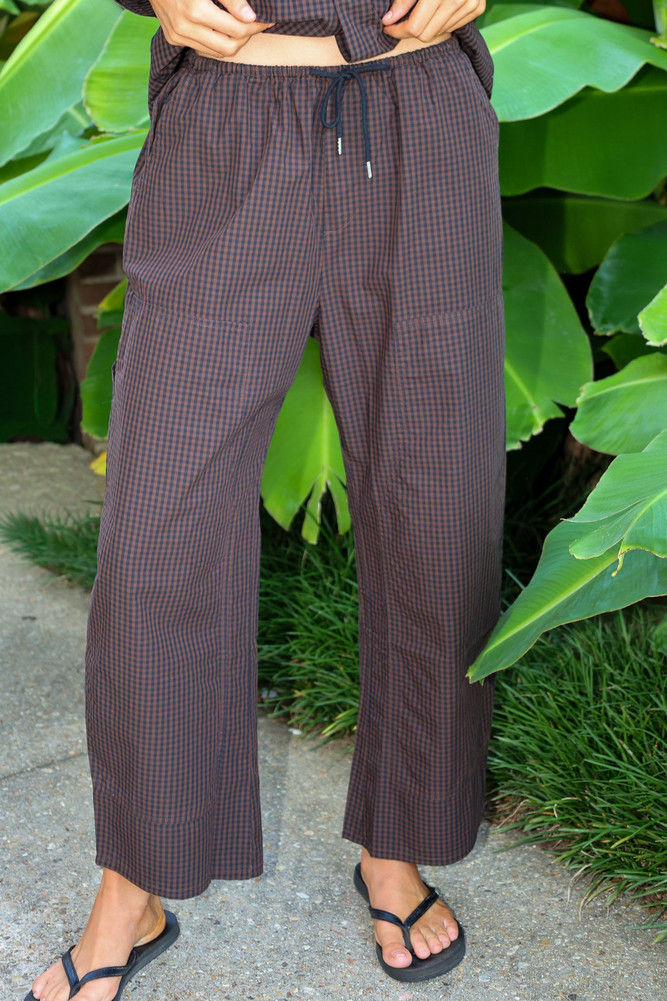 A person wearing loose, wide-leg cropped barrel pants by PISTOLA in a dark brown and black micro-check pattern, standing outdoors on a stone surface with green tropical leaves in the background.