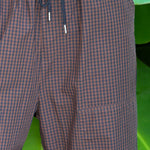 Close-up of a person wearing brown and black gingham check cargo pants with a dark drawstring, set against a background of large green tropical leaves.