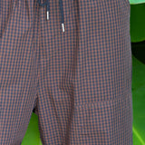 Close-up of a person wearing brown and black gingham check cargo pants with a dark drawstring, set against a background of large green tropical leaves.