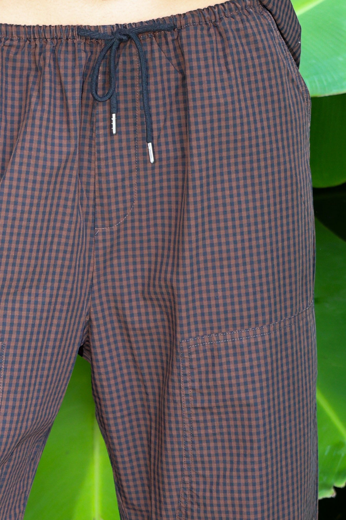 Close-up of a person wearing brown and black gingham check cargo pants with a dark drawstring, set against a background of large green tropical leaves.