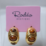 A pair of gold hoop earrings with three brown tiger's eye stones each, displayed on a pink Rodéo Boutique card.