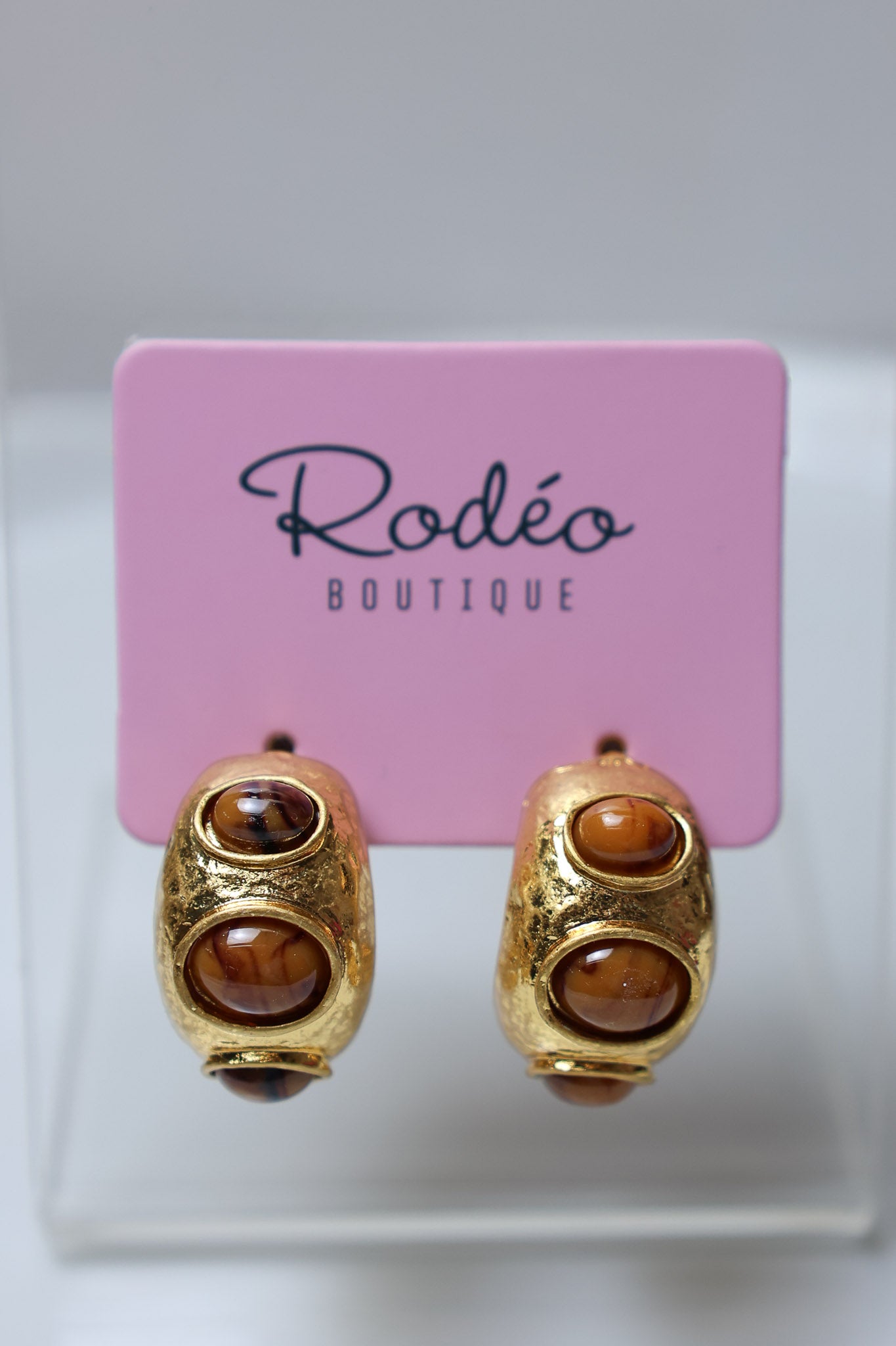A pair of gold hoop earrings with three brown tiger's eye stones each, displayed on a pink Rodéo Boutique card.