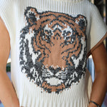 A close-up of a woman wearing a cream ribbed knit sweater with a large knitted tiger face graphic on the front in beige, brown, and black.