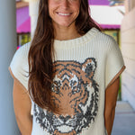 A smiling woman with long brown hair wearing a cream ribbed knit sweater with a large tiger face graphic in black, gray, and tan on the front, paired with light gray jeans.