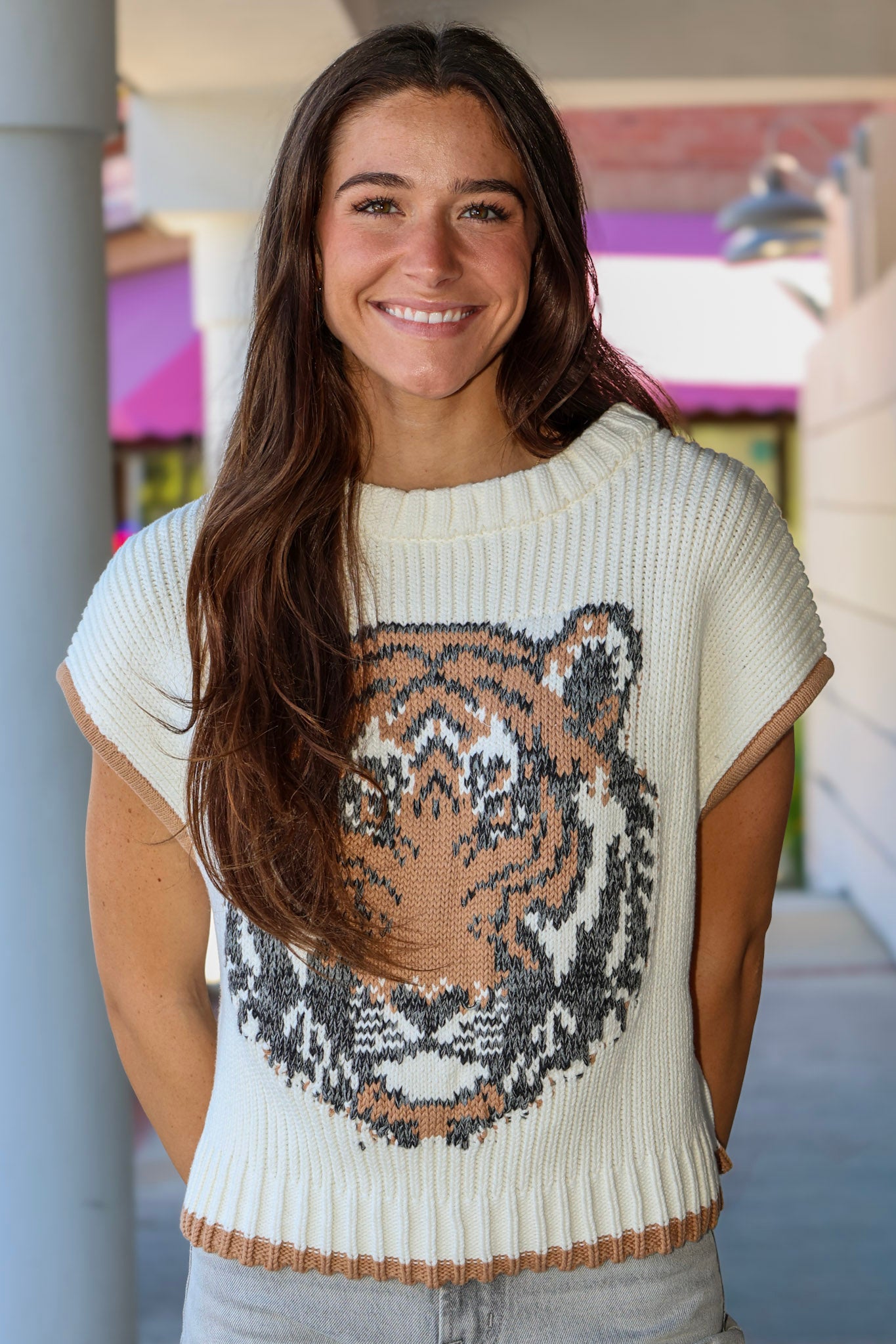 A smiling woman with long brown hair wearing a cream ribbed knit sweater with a large tiger face graphic in black, gray, and tan on the front, paired with light gray jeans.