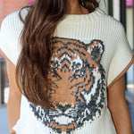 A woman wearing a cream-colored short-sleeve knit sweater featuring a large knitted tiger face graphic on the front, paired with light blue jeans.