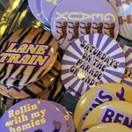 A gold-colored tray filled with various round pinback buttons featuring purple, yellow, black, and white designs, including tiger graphics and text such as 'LANE TRAIN', 'TIGER TERRITORY', and 'HEAUX'.