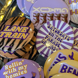 A gold-colored tray filled with various round pinback buttons featuring purple, yellow, black, and white designs, including tiger graphics and text such as 'LANE TRAIN', 'TIGER TERRITORY', and 'HEAUX'.