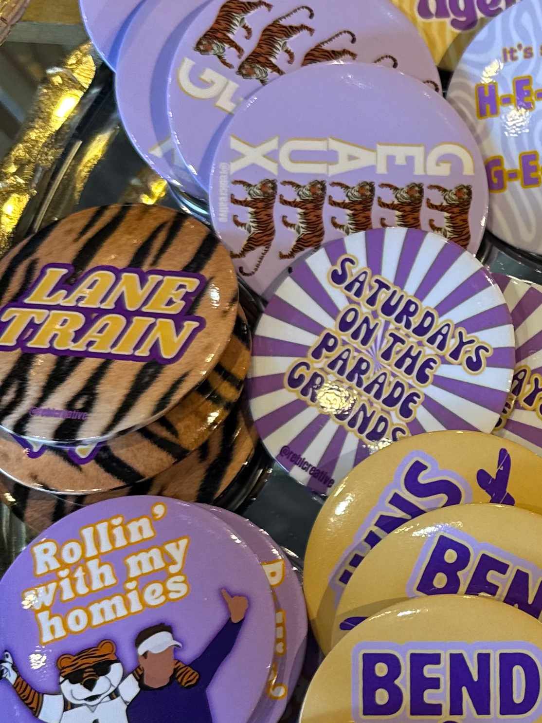 A gold-colored tray filled with various round pinback buttons featuring purple, yellow, black, and white designs, including tiger graphics and text such as 'LANE TRAIN', 'TIGER TERRITORY', and 'HEAUX'.