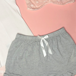 Collection of women's clothing including a white tank top, gray shorts, lace panties, and socks on a pink background.
