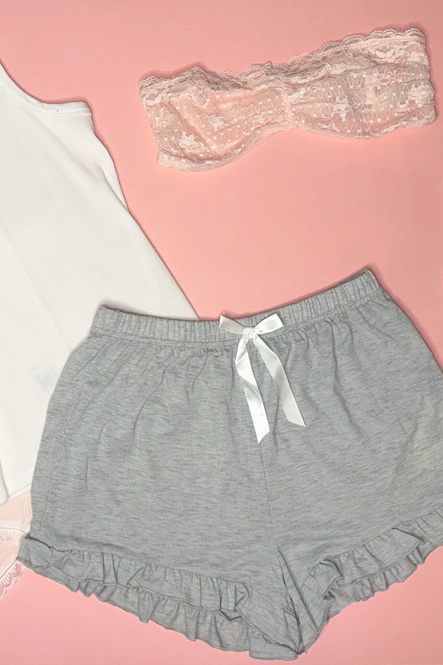 Collection of women's clothing including a white tank top, gray shorts, lace panties, and socks on a pink background.