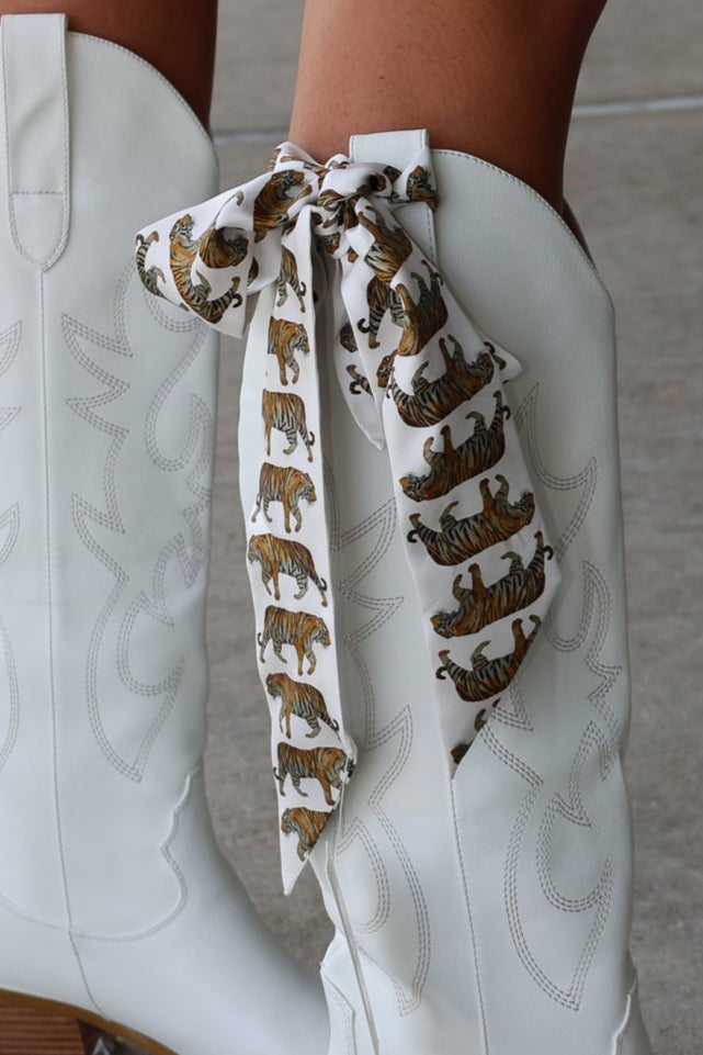 White cowboy boots with a decorative tiger print ribbon on a neutral background