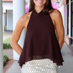 A woman modeling a dark brown halter-neck top paired with a white sequined mini skirt, standing in front of a modern building with white columns.