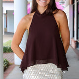 A woman modeling a dark brown halter-neck top paired with a white sequined mini skirt, standing in front of a modern building with white columns.