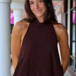 A woman wearing a sleeveless high-neck top in dark brown with a sheer hem, paired with a white sequined skirt, standing in front of a building with white columns.