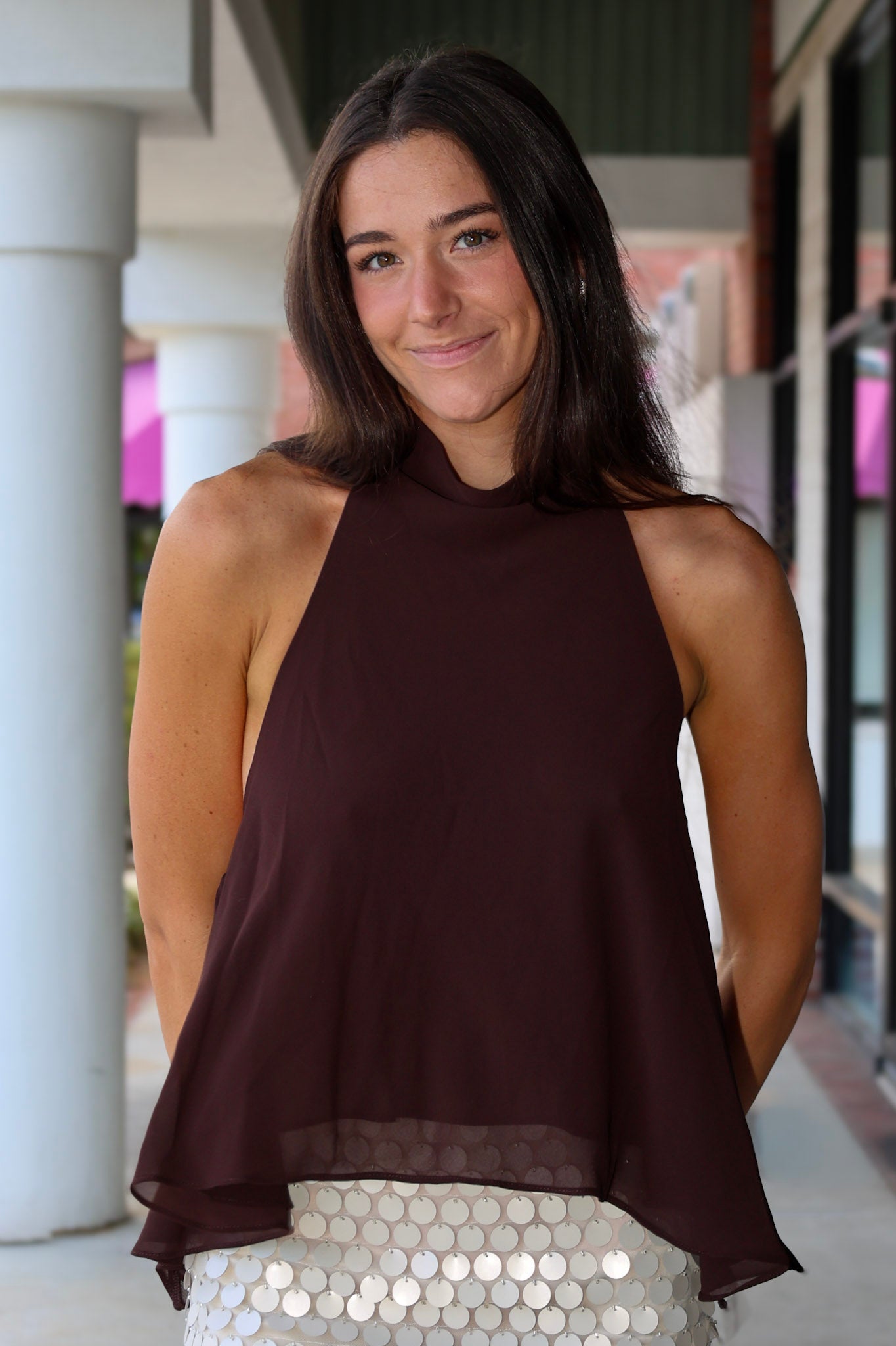 A woman wearing a sleeveless high-neck top in dark brown with a sheer hem, paired with a white sequined skirt, standing in front of a building with white columns.
