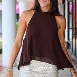 A woman modeling a sleeveless high-neck halter top in dark brown, paired with a white sequined mini skirt, standing in front of a building with white columns.