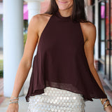 A woman modeling a sleeveless high-neck halter top in dark brown, paired with a white sequined mini skirt, standing in front of a building with white columns.