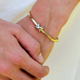 A close-up of a person's hands and wrists, showcasing a delicate gold bangle bracelet with a small silver-toned butterfly or cross-shaped charm on the wrist, alongside a silver-toned ring on the finger, set against a soft white fabric background.