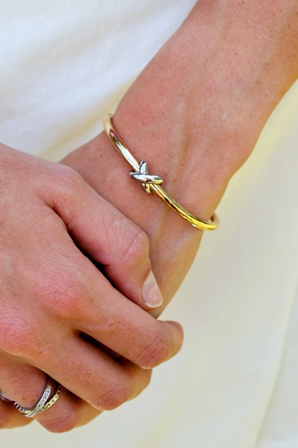 A close-up of a person's hands and wrists, showcasing a delicate gold bangle bracelet with a small silver-toned butterfly or cross-shaped charm on the wrist, alongside a silver-toned ring on the finger, set against a soft white fabric background.