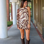 A woman modeling a one-shoulder mini dress with a brown and beige tiger print, paired with dark brown cowboy boots, walking along a covered outdoor walkway near Rodéo Boutique.