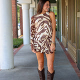 A woman modeling a one-shoulder mini dress with a brown and beige tiger print, paired with dark brown cowboy boots, walking along a covered outdoor walkway near Rodéo Boutique.
