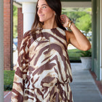 A woman modeling a one-shoulder mini dress with a brown and beige tiger print, paired with dark brown cowboy boots, walking along a covered outdoor walkway near Rodéo Boutique.