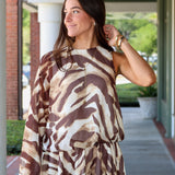 A woman modeling a one-shoulder mini dress with a brown and beige tiger print, paired with dark brown cowboy boots, walking along a covered outdoor walkway near Rodéo Boutique.