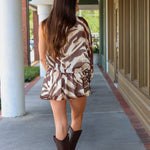 A woman modeling a one-shoulder mini dress with a brown and beige tiger print, paired with dark brown cowboy boots, walking along a covered outdoor walkway near Rodéo Boutique.